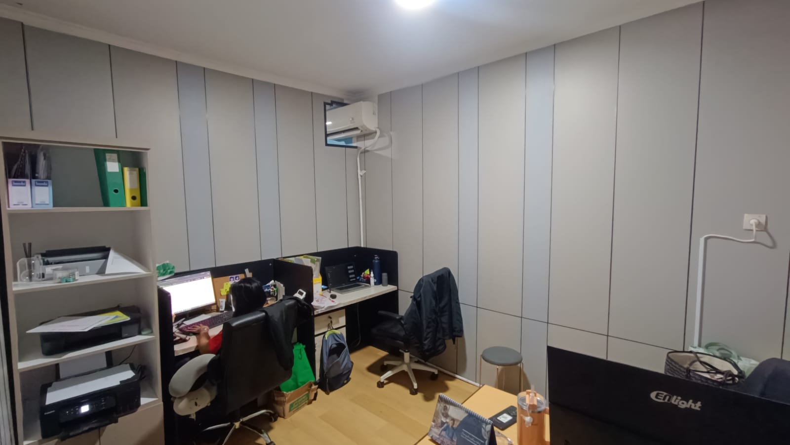 Ruang kerja dengan meja, komputer, rak dokumen, dan seorang staf.