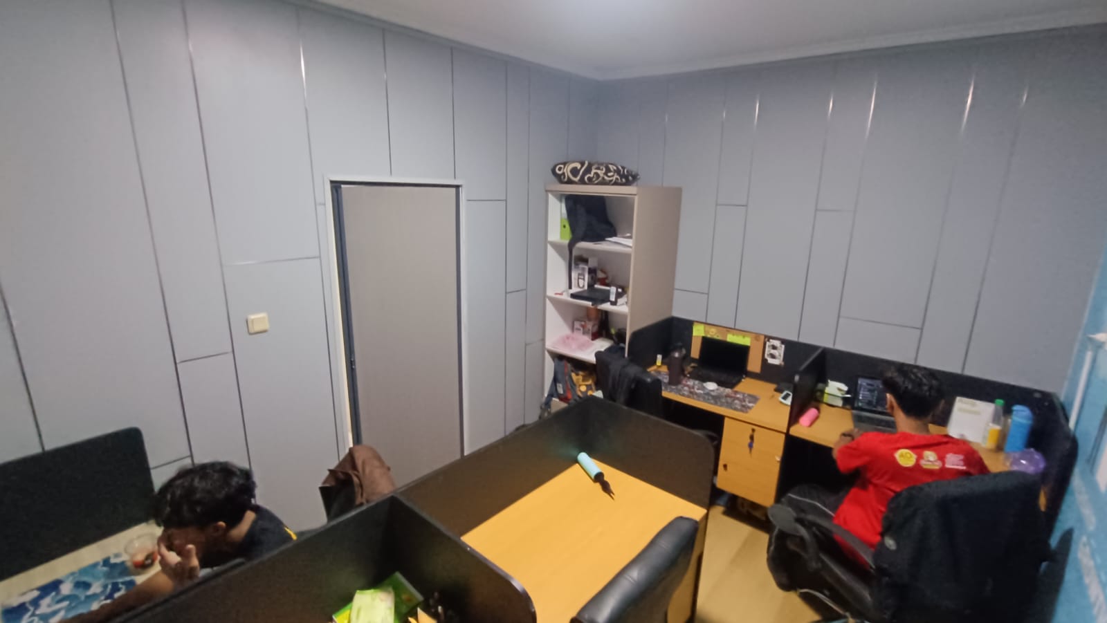 Ruang kerja dengan meja, komputer, rak, dan dua orang.
