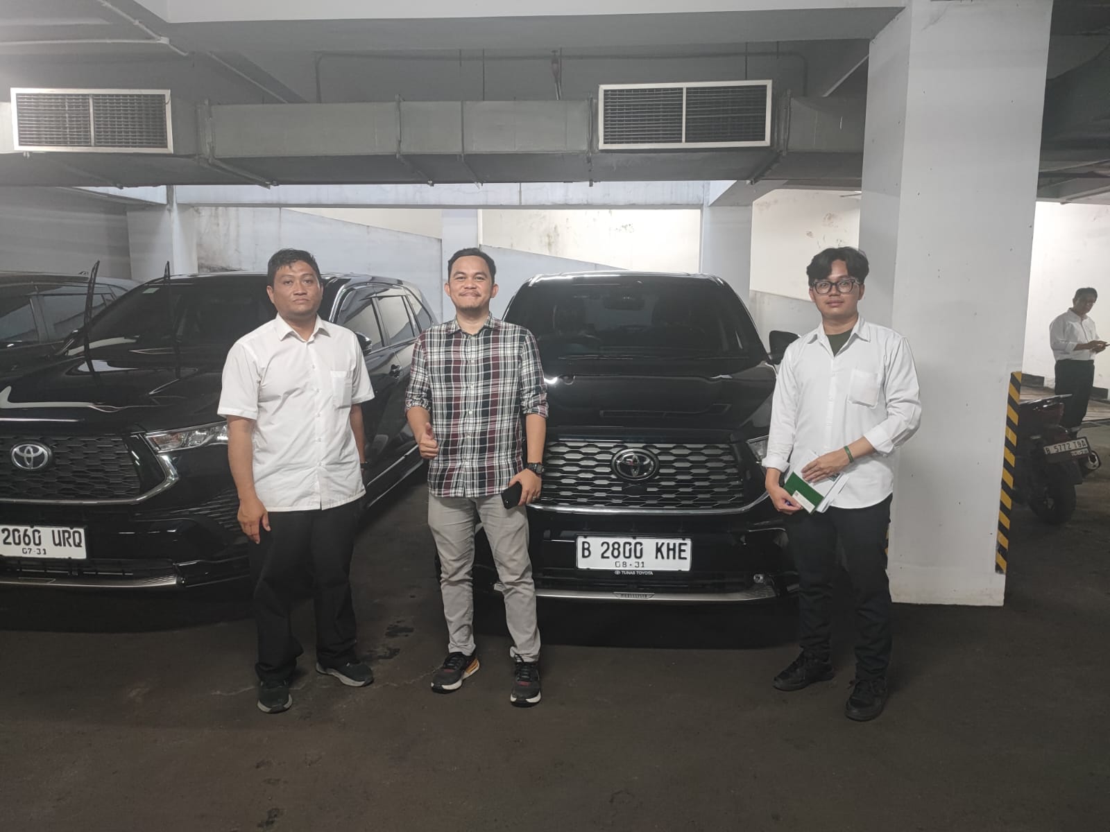 Tiga orang berdiri di depan dua kendaraan hitam di area parkir.