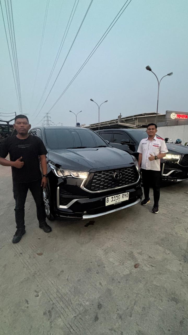 Dua orang berdiri di depan kendaraan hitam dengan kendaraan lain di belakang.